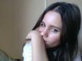 NellaKoczur has long dark brown hair, brown eyes, and is covering her nose with her hand, looking shy. She's wearing a white shirt and has a slight blush.
