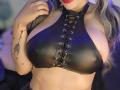 Kkole's large, shiny black leather top with lace-up front, highlighting her big, round tits. She has a tattoo on her left arm and red lipstick.