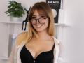 ChloeKingsley with blonde hair, black glasses, and a smile, wearing a black lace bra and sheer white shirt, exposing her tits. She's cute and has a sexy look.