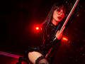 TatsumiLee with black latex outfit, thigh-high lace stockings, and glasses, holding a pole, red lighting, dark makeup, and black lipstick, posing seductively, showing her ass.