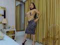 PamelaLiss with long black hair, wearing a leopard print skirt, black high heels, and a strapless top revealing her tits, standing in a bedroom with yellow curtains.