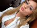 EliseMonet with pouty lips, glasses, blonde hair, white shirt, gold necklace, showing cleavage, small gold earrings, looking at camera, wooden background, selfie, medium breast size.