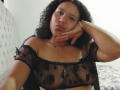 AriaVega with curly black hair, pouting lips, and hand on cheek, wearing a sheer black top with black polka dots, showing her small tits and a delicate necklace.
