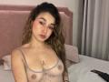 MilaGonz with wavy brown hair, pink eyeshadow, and a cute face, wearing a sheer gray top, exposing her tits and a flower tattoo below. She's on a bed.
