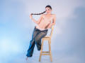 AnabelleRoss is topless with small tits, holding a long brown braid, wearing blue jeans and white panties, sitting on a wooden stool, barefoot, with a tattoo on her left arm.