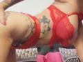 FrenchShanna in red lace lingerie, showing her tits and tattooed side with a cat face, leaning forward with a hand on her hip and red nails.