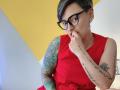 KatParadise with short black hair, blue glasses, red dress, tattooed arms, and finger in mouth, looking cute and thoughtful. Her tits are small, and she's sexy as hell.