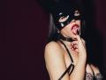 JaneWow with black mask, red lipstick, and finger in mouth, touching tongue to finger, wearing black strap lingerie, showing cleavage, and black choker, against dark background.