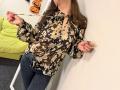Tilliana vestindo uma blusa floral marrom, calças jeans escuras, com unhas cor-de-rosa, segurando uma corda, mostrando os seios, em pé em frente a uma parede branca, com um sofá verde ao fundo e decorações de Halloween na parede..