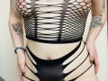 KarKutie wearing a black fishnet top with large holes exposing her tits and a matching black thong with strappy sides, showing tattoos on her arms and a bracelet.