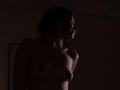 Darkly lit photo of MaryCruzz with short curly hair, small tits, and a tattoo on her left shoulder, standing side profile, shadowing her upper body.