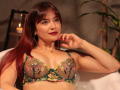 KateBurning with red hair, wearing a green and yellow floral lace bra, showing her tits, red lipstick, and a necklace, hand near her mouth, candle in the background.