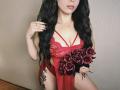 Niferttiti with long black hair, wearing red lace lingerie, holding a bouquet of black roses, showing her tits, and a thigh tattoo, has black nails and a necklace.