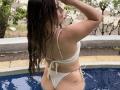 LorensDouce with wet brown hair, white bikini, showing ass, small heart tattoo on right buttcheek, standing in blue pool, arms raised.