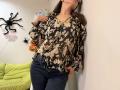 Tilliana, vestindo uma blusa florida que mostra seus seios, encostada em uma parede, usando jeans azuis, com óculos, e com a mão dentro da calça, em um quarto com tema de Halloween..