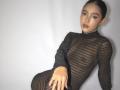 GabySmiith with a sleek, dark, sheer dress, showing her small tits through the fabric, dark nails, and a confident, sexy pose, against a plain white background.