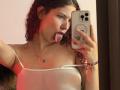 ValeryScot with curly red hair, tongue out, wearing a white spaghetti-strap top, holding a phone, red light on her. Boobs visible through top, necklace, bracelet. Mirror selfie.