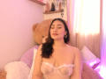 ElyRoss with closed eyes, wearing a sheer white bra that exposes her tits, has dark brown hair, and gold hoop earrings, sitting in front of a giant teddy bear.