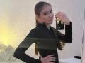 DomingaSierra with long brown hair in a ponytail, wearing a black long-sleeve top, taking a mirror selfie, hand on hip, slight smile, holding a phone with a gold case.