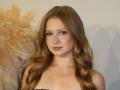 AkilahGhil with long, wavy blonde hair, blue eyes, and nude makeup, has a cute face and small, perky tits, standing against a plain background with feathery decor.