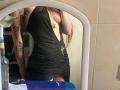 Mettis in a black, deep-cut dress showing tits, with tattoos on arms, wearing a chain belt, reflected in a mirror, long brown hair, bottle in the background.