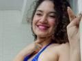 SanetPerlas, curly-haired, smiling with pink lipstick, wearing a blue halter top, pointing with right finger, showing her tits, standing in a white-tiled bathroom.