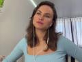 TangelaEmmel has medium brown hair, wearing a light blue top, with white earbuds in, pink lipstick, and a slight pout, looking off-camera. Her tits are slightly visible.