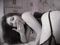 Aeron69 with long brown hair, black lace lingerie, and garter belt, on bed, bent over, hand near mouth, looking at camera, showing her ass and fishnet stockings.