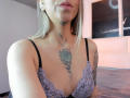 KathyM with blonde hair, wearing a lace bra, showing a rose tattoo on her chest and a star tattoo on her neck, standing in a softly lit room.