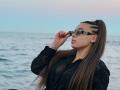 Niferttiti with long brown hair in braids, wearing black sunglasses, black patterned shirt, and red nails, adjusting her sunglasses, standing by the ocean.