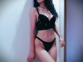 SweetClaraa with curly black hair, wearing a black lace bra and matching panties, standing against a white wall, showing her tits and pussy. Her skin is light, and she's wet.