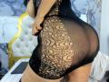 NaomyHott69 in a black, see-through lace dress, showing her big ass and fat thighs. Her hand rests on her side. Laptop on the bed.