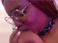 RoseBeckers with red hair, wearing glasses, sucks on a large, dark nipple with a shiny black collar, purple lighting. Her finger is near her lips, looking focused.
