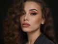 Niferttiti has wavy brown hair, full lips with nude lipstick, and smoky eye makeup. She's looking serious with a dark background, wearing a black top.
