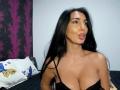 BrunetteBabe69 with long black hair, pouty lips, and large tits in a black lace top, looking to the side, in a bedroom with a purple-patterned wall.