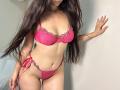 Tisia with long brown hair, wearing a pink lace bra with floral details and matching pink lace panties with side ties, standing against a white wall.