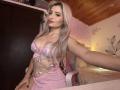 EliseMonet, avec de longs cheveux blonds, en lingerie en dentelle rose qui révèle ses seins, assise sur une chaise rose, prenant un selfie, plafond en bois, lumière chaude dans la pièce, sourire discret, pose détendue..