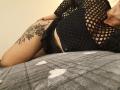 Niferttiti's black fishnet top reveals her large tits, and she's lying on a grey patterned blanket, showing a thigh tattoo and black panties. Her skin is light.