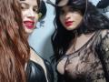 Two HotSexyBigCum females with red lipstick, one with brown hair and black leather top, the other with black hair and see-through lace top, showing tits.