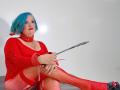 LeidiBlue with blue hair, red lipstick, and red outfit, wearing a collar and cuffs, holding a strap, sitting with fishnet legs, showing her tits.