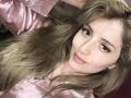 EliseMonet with long brown hair, wearing a pink satin shirt, smiling at the camera, slightly tilted head, brown eyes, subtle makeup, small gold earrings, soft pink lipstick.