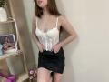 StellaNoir with small tits, wearing a white lace corset and black satin skirt, hands on skirt, standing in a room, brown hair, cross necklace, light skin.