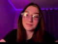 SunnyKlusman with brown hair, glasses, nose ring, smiling slightly, wearing black shirt, purple LED lights in background, looking into camera, cute, nerdy.