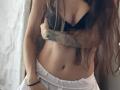 VelvetRay con lunghi capelli castani, reggiseno nero e pantaloni bianchi, braccia incrociate sul petto, tatuaggio sul braccio destro, braccialetto nero al polso sinistro, luce soffusa..