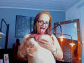 SamantaMature69 with glasses, tongue out, grabbing her large, bare tits with both hands, pink shirt lifted, in a brightly lit room with wall sconces.