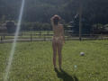 AlexXas stands with back to camera, wearing a thong and topless, sunlight flares to left, grassy field, wooden fence, small house in background, holding hair up.
