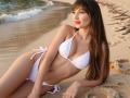 Rouzi, con cabello largo y castaño, traje de baño blanco y lápiz labial rojo, acostada en una playa arenosa, mostrando sus pequeños senos y su delgada figura, mirando a la cámara con una ligera sonrisa..