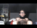 LapijArgie69 with glasses, shirtless, black cloth around neck, hairy chest, sitting on bed, legs up, looking at camera, room in background, dark wall behind.