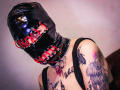AmySpider with shiny black latex mask, red lace-up front, wearing black tank top, showing tattooed arms and chest, silver chain collar, and red pendant, in a provocative pose.