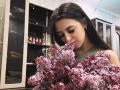 BokSinha with dark brown hair, green top, holding a big bunch of pink lilacs, smelling them, smiling slightly, in a kitchen with wooden cabinets and glass doors.